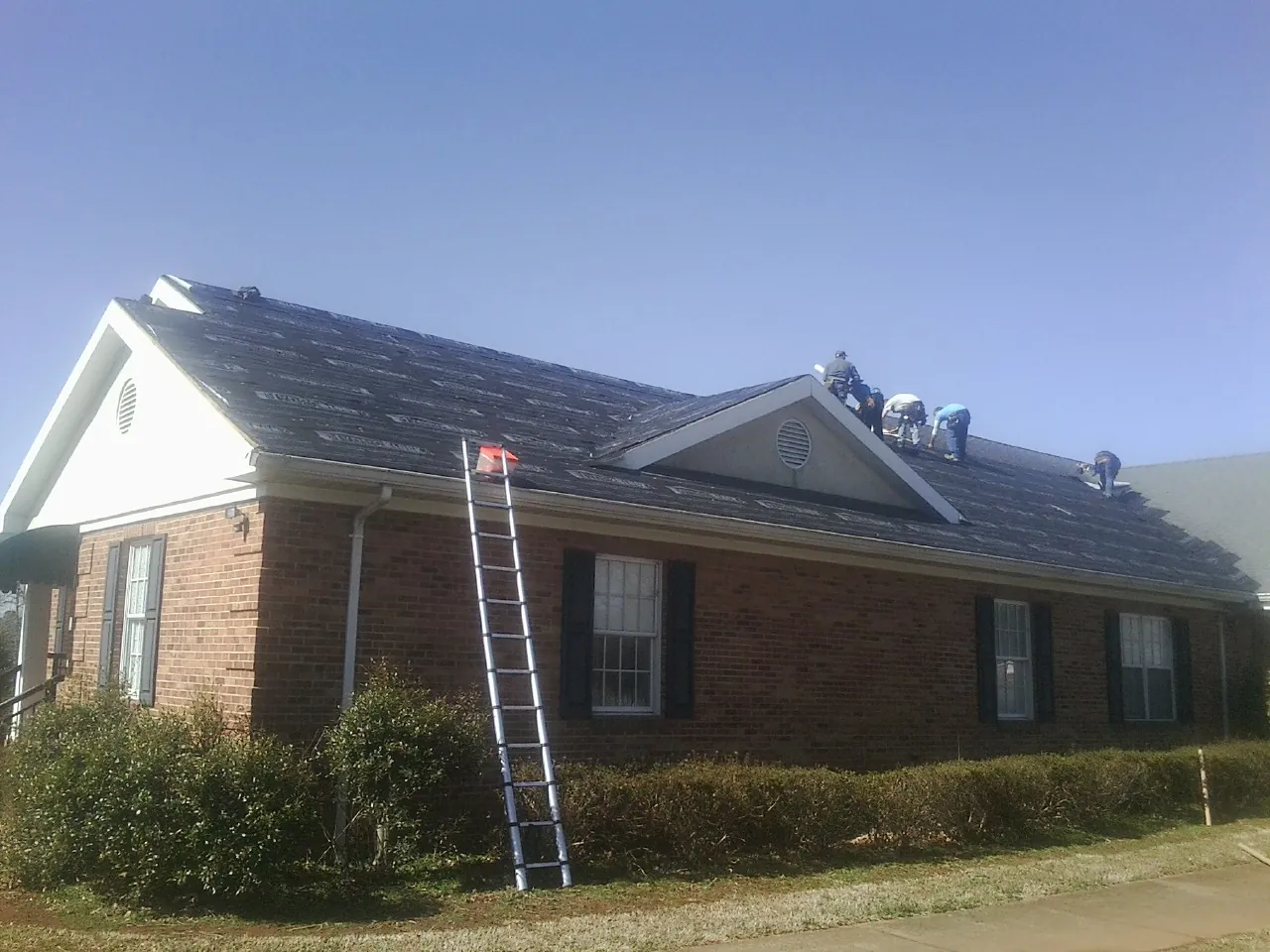 Crew tearing shingles off a brick ranch roof with a ladder set — Emergency Roof Repair work by our crew in Aldrich