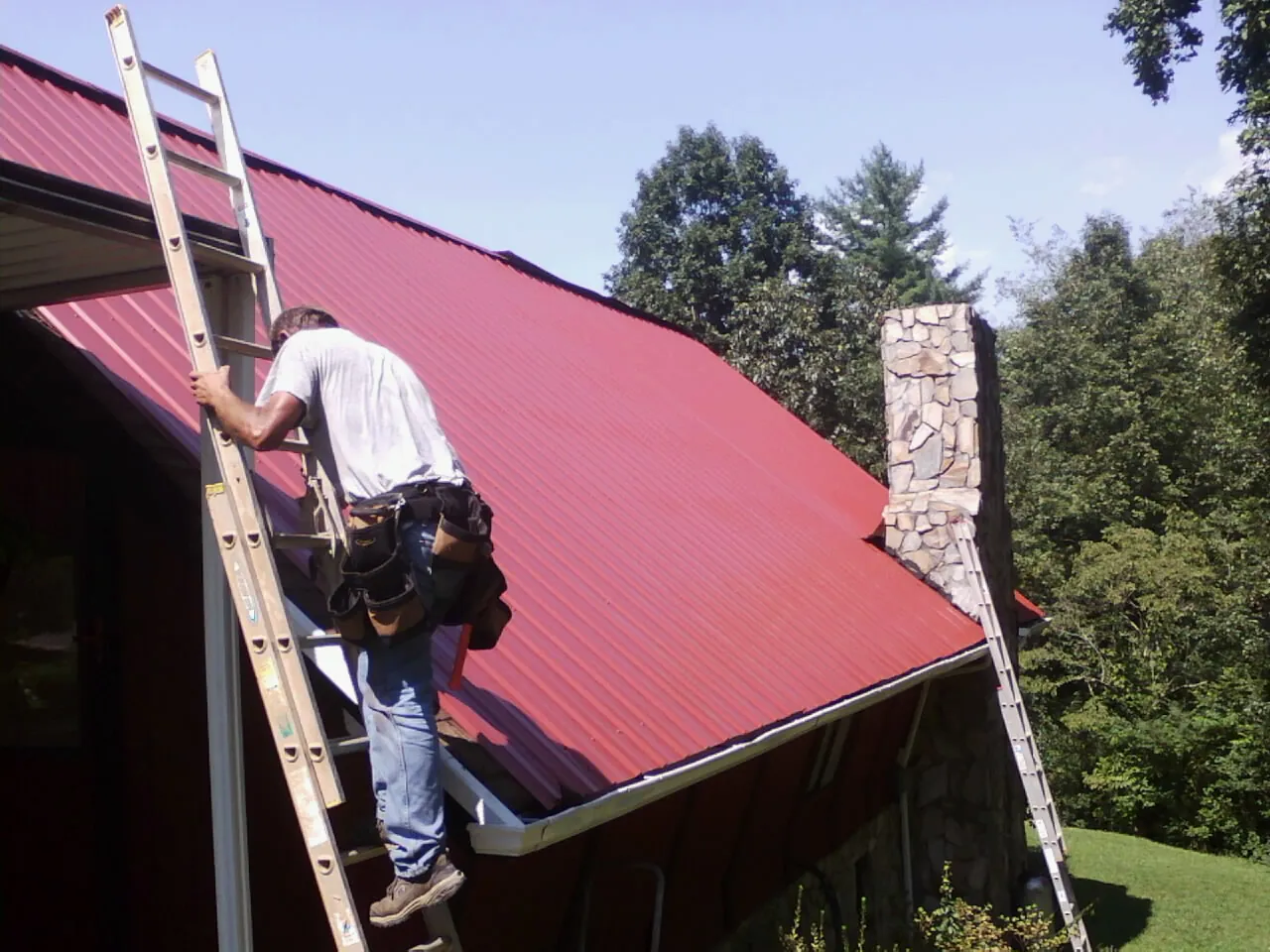 Roofer on a ladder beside a red metal roof and stone chimney — Soffit & Fascia Repair by Roofing Brothers in Aldrich