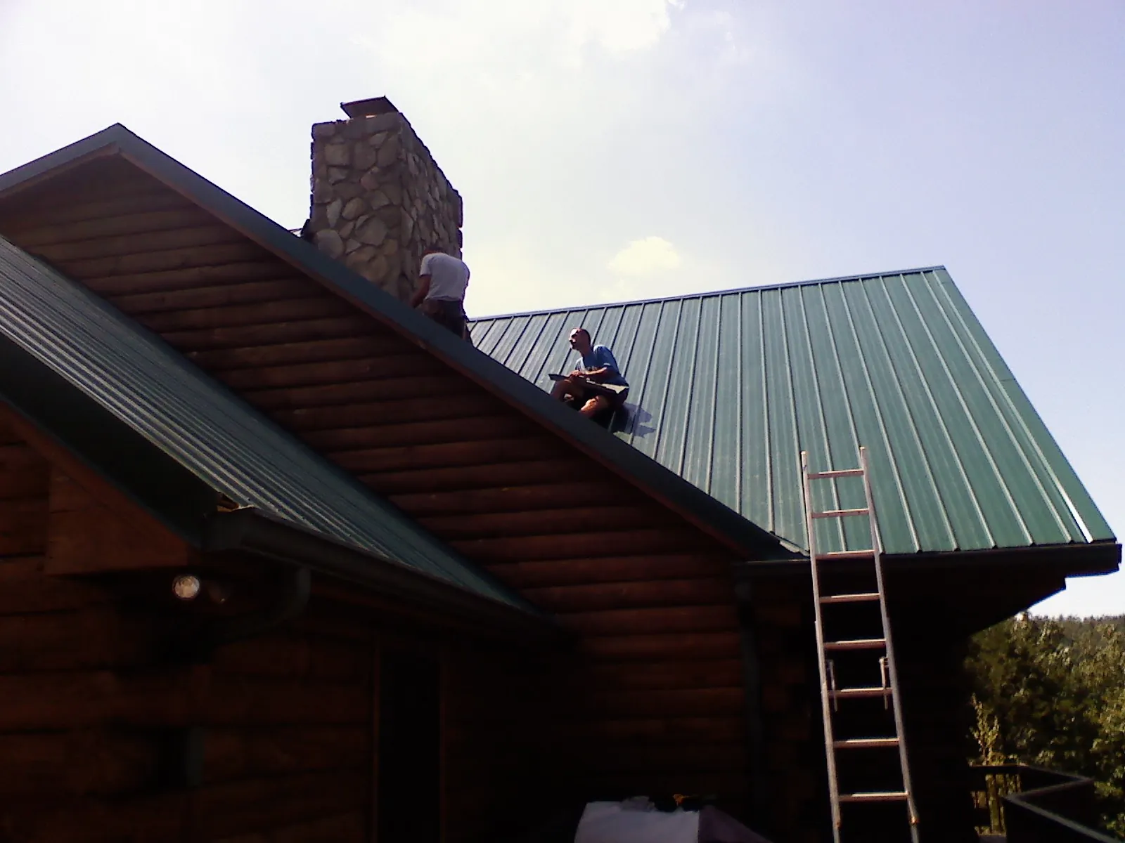 Roofers installing green metal panels beside a stone chimney on a log home in Aldrich